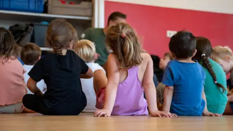 Getty Images Children sitting on the floor attentively during story time, with their backs to the camera.