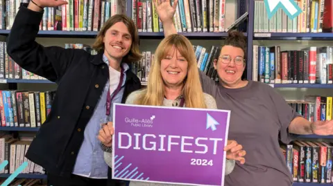 Guille-Allès Library Three people stood in front of three bookshelves side by side. The person on the left is wearing a blue shirt with a white vest underneath and a blue jacket. The person in the middle is wearing a grey cardigan and is holding up a sign that says DIGIFEST 2024 in white writing on a purple background. The person on the right is wearing a grey t-shirt. All three people are smiling and the people on the left and right have their hands up in excitement. 