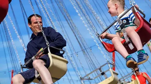 Family A man wearing navy shorts and a coat riding a fairground ride looking at a little boy wearing a striped top.