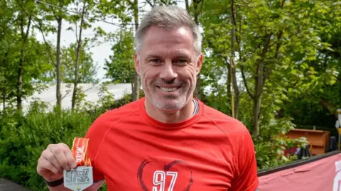 Paul Francis Cooper Former Liverpool footballer Jamie Carragher wearing a red BTR Run For The 97 sports top on smiles as he holds a red and silver 97 5K medal up on a sunny day.