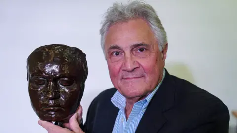 Getty Images A man with grey hair, a dark suit and striped open-necked shirt holding a model of a persons face