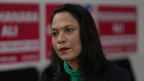 Rushanara Ali wearing a black blazer over a green rollneck top. She has long black hair and has a serious expression on her face while sat in an office.