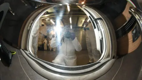 COPFS A stainless steel teapot. The reflection shows a man taking a photograph of the teapot. 