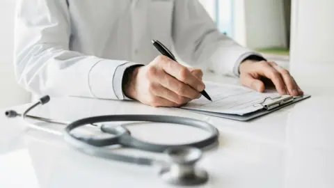 A man wearing a white lab coat is sitting at a white table filling in a form on a clipboard with a black pen in his right hand. In the foreground, also sitting on the table, is a stethoscope. 