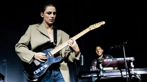 Ellie Rowsell of Wolf Alice plays a black Fender telecaster guitar while wearing a beige blazer and necklace. A man in the background smiles while sitting in front of a Roland keyboard