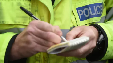A close up of a police officers hands and chest as he makes notes with a pen on a small notepad. He is wearing a yellow hi-vis jacket with "Police" on it and is wearing a watch.