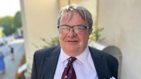 Deputy John Gollop has grey and black hair and is looking at the camera. He is wearing black rectangle rimmed glasses and smiling. His suit is black and he is wearing a white shirt with a purple tie with small Guernsey flags on it. 
