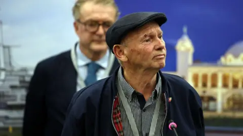 Bryan Macdonald looking to the right. He is wearing a dark, flat cap and a dark striped shirt with a navy blue jacket over the top. There is a man standing out-of-focus behind him.