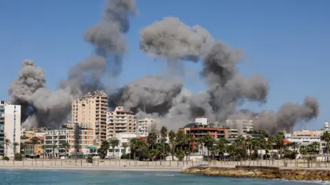 Reuters A fumaça sobe após os ataques israelenses à cidade de Tiro, no sul do Líbano (23 de outubro de 2024) - A costa de Tiro é visível da água, vários prédios altos de tamanho médio e árvores podem ser vistos na praia de Tiro.