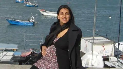 Geeta is heavily pregnant, sat outdoors with the coast behind her and small boats on the water. She is wearing a floral long top, with black trousers, cardigan and jacket and has her hand on her bump.