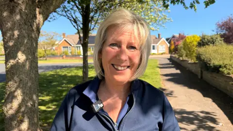 Jasmine Lowe / BBC Irene Hay - a woman with a blonde bob smiling at the camera. She is standing in a rural village under a large tree in the shade. She is wearing a navy blue quarter zip top.