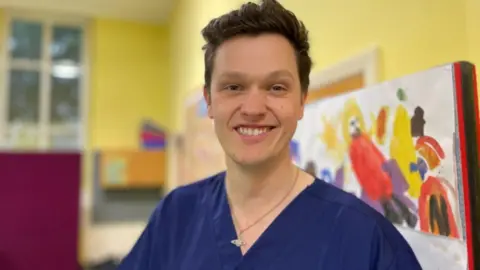 Ross Keat, a man with brown hair and a bright smile, he wears a blue dental uniform and is stood in a school.