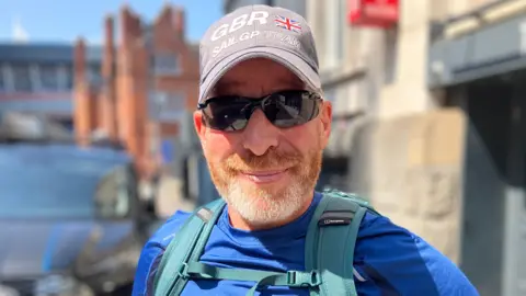 Paul is wearing a grey baseball cap and dark sunglasses, with a blue t-shirt, and is looking into the camera in Cardiff City centre. He has grey and ginger stubble and wears a light blue backpack. 