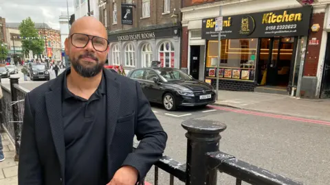 Amy Holmes/BBC Matthew Bushnell stood in Luton town centre. He is leaning against a black fence and is wearing a black jacket and black polo shirt. He has glasses and a black beard and moustache.