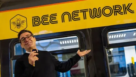 TFGM Andy Burnham, in a black jumper holding a microphone, stands in front of a yellow Bee Network vehicle
