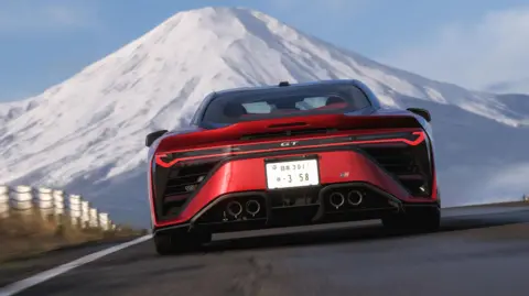 Screenshot shows the back of a red sports car as it races along a mountain road towards the snow-capped peak of Mt Fuji.