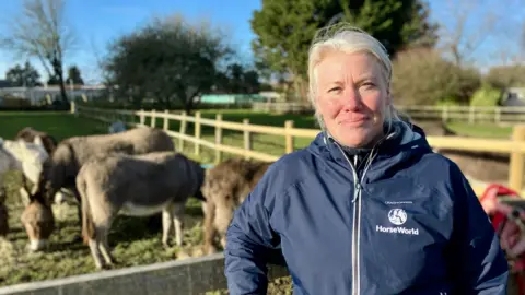 Sara smiles at the camera in front of a paddock with a number of grey donkeys in it. She has very bright blonde hair and wears a navy HorseWorld raincoat which is zipped up.