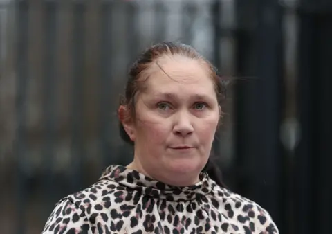 Pacemaker A woman, standing in front of a beige building, looking at the camera. She has brown hair, tied back. She is wearing a leopard print top. 