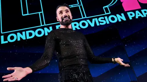 Rylan Clark on stage at London Eurovision Party - he wears a sparkly black top and his arms are open