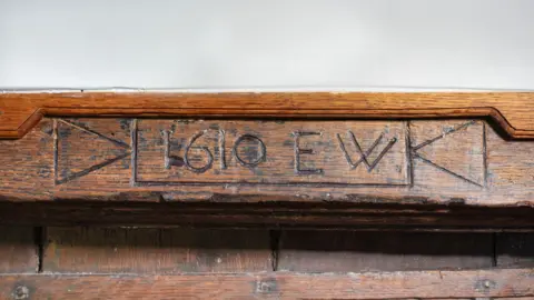 Historic England Archive A close-up of a brown wood moulded lintel which bears the date 1610 and the initials E.W above a door. 