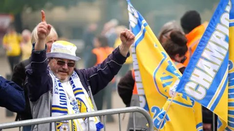 Man in Leeds hat and scarf holds up his hands in celebration, with big flags to one side 