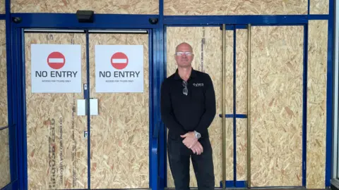 Steve Tyler stands in front of a doorway with boards on