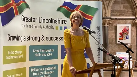 Dame Andrea Jenkyns with blond hair and wearing a yellow dress in front of a banner for the Greater Lincolnshire Combined County Authority