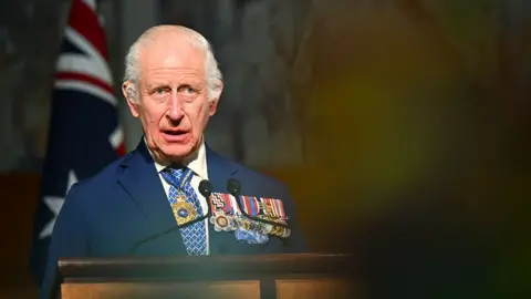 EPA King Charles delivers a speech in the Australian Parliament on Monday, he is wearing a suit and white shirt with a blue and white tie, with several medals on his left lapel and a gold necklace around his neck. He's looking slightly away from camera, with an out-of-focus audience member in the foreground and an Australian flag in the background.