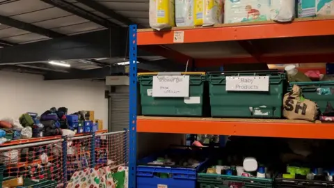 A warehouse with crates of donated goods - 3 shelves filled with crates of shower gels, baby products, shampoos and other cleaning items are pictured.