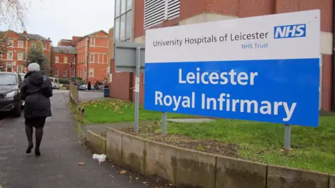 Leicester Royal Infirmary sign outside entrance with woman in hat walking past