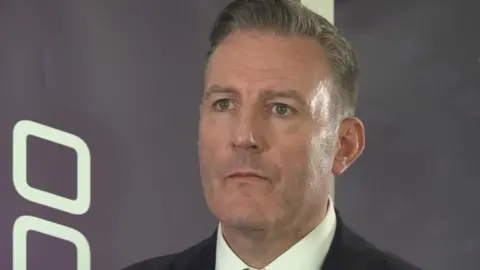 Clive Higgins - a man wearing a suit, standing in front of a purple background. He has grey/brown hair.