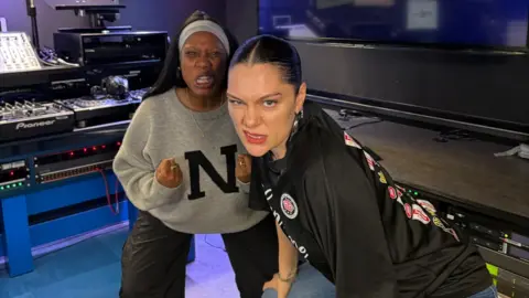 Jessie J and Nadia Jae make faces while crouching in a radio studio