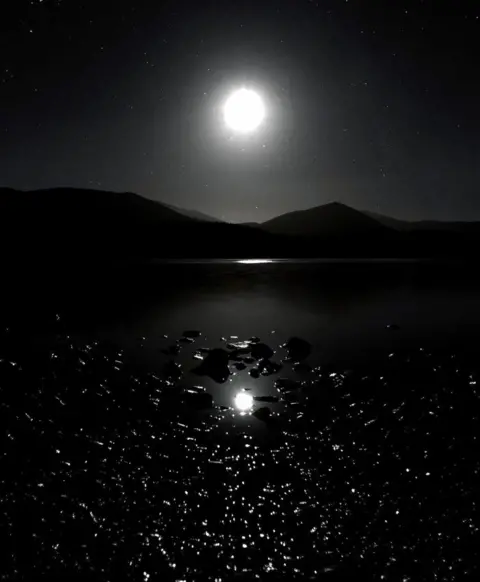 @peter_diender Une photographie du Loch Morlich dans le parc national des Cairngorms dans l'obscurité. Il montre la lune dans le ciel nocturne se reflétant sur l'eau en dessous 