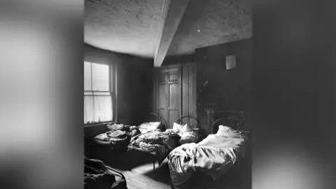 Image courtesy of Manchester Libraries A black and white photograph of a common lodging house showing four beds with metal frames in a dark and bare room. It is dated as 1897 and was a room at 44 Angel Street, Angel Meadow, Manchester.