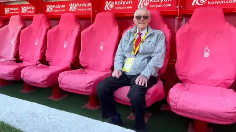Alan can be seen sitting in the manager's dugout and smiling to camera