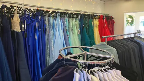 BBC/ Joanita Musisi A formalwear boutique with racks of dresses arranged in a colour gradient from dark blues to bright reds, alongside a circular rack of men’s suits and shirts, all set against a backdrop of warm string lights.