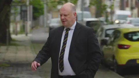 A bald white man in a dark suit and light shirt and dark tie walking up across a pavement by some cars