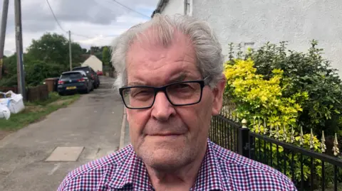 Neville Patrick looks directly at the camera wearing black glasses and a chequered shirt. He is photographed outside with residential houses behind him and a pathway with two cars parked on the right hand side. 