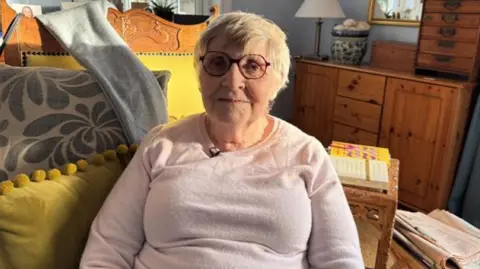 BBC Anne Campbell is an older woman. She is short white hair and is wearing glasses and a light pink jumper. Her room is furnished with a wooden dresser and cushions.