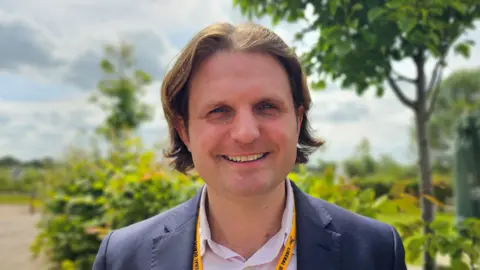Paul Moseley/BBC Steffan Aquarone standing in front of green foliage outdoors. He is wearing a blue blazer with a yellow lanyard round his neck. He is looking directly at the camera and smiling.