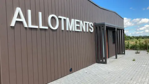 Emma Howgego/BBC A brown-panelled building with the word "Allotments" written on the front. There is a grey block paved area to the front with greenery beyond that. 