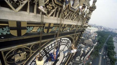 A fisheye lens view of the top of the clock tower of Big Ben. Looking down towards the top of the clock face, Peter Duncan and his cameraman and one of the Steeplejacks are seen hanging off the top of the clock tower on ropes.  Buildings, trees and a road are visible on the right hand side , that gradually disappear into the horizon.