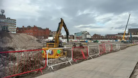 A yellow digger is on a construction site behind red and white fencing and a concrete path.