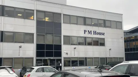 The image shows a modern, three‑storey office building with a mostly white and grey exterior. Large rectangular windows line each floor, with some panels appearing darker than others, giving the façade a slightly varied pattern. The building has the name “PM House” displayed prominently in dark blue lettering on the front.
Several cars are parked in the foreground.