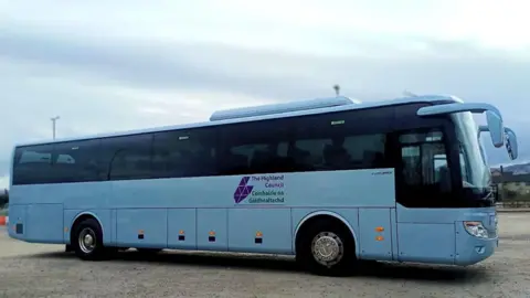 A bus in Highland Council white and purple colours is parked on a large area of hard standing gravel. Some houses and lampposts can be seen in the background. 