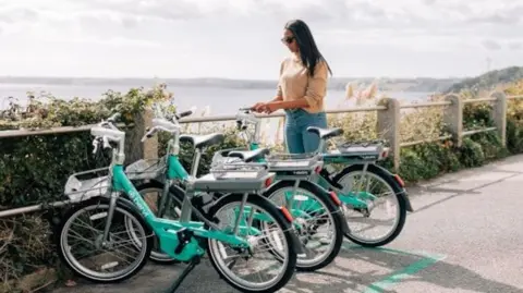 Dorset Council Three light green bikes standing up aside a path overlooking the coast, with a woman with sunglasses stood next to one, trying to get it started 