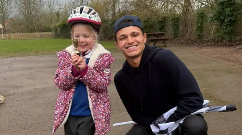 Chew Stoke Church School Lando is smiling in a photo kneeling next to a little girl wearing a bike helmet. The girl is holding a mini trophy and smiling with excitement. They are both standing in the school playground.