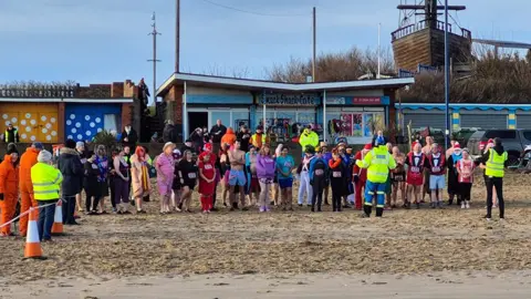 Carl Tebbutt & Diane Martin Dozens of swimmers, some in fancy dress, stand on the beach at Mablethorpe with safety marshals in fluorescent jackets addressing the crowd