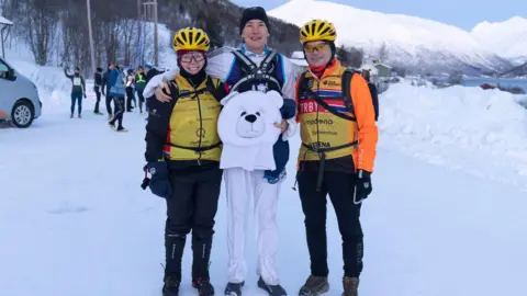 Gill Punt Three people standing on ice and snow in Norway. The woman is the middle is wearing a polar bear costume and holding the head, the other two people are dressed in orange and yellow. They are smiling at the camera. 