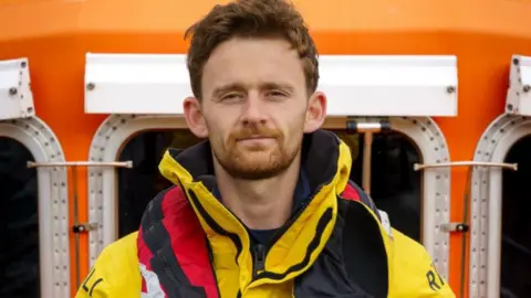 A man with ginger-coloured hair and beard wearing a yellow coat and a red life jacket.